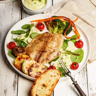 Fried sea fish and vegetables. Tilapia fillet with broccoli, cherry tomatoes and lettuce. Sea food on a white plate on white background. Vertical shot